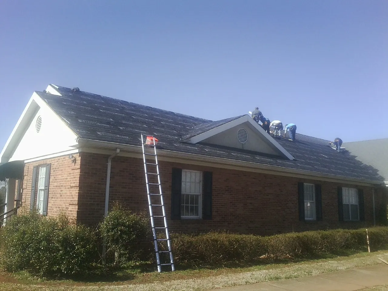 Crew tearing shingles off a brick ranch roof with a ladder set — Emergency Roof Repair work by our crew in Michigan Center
