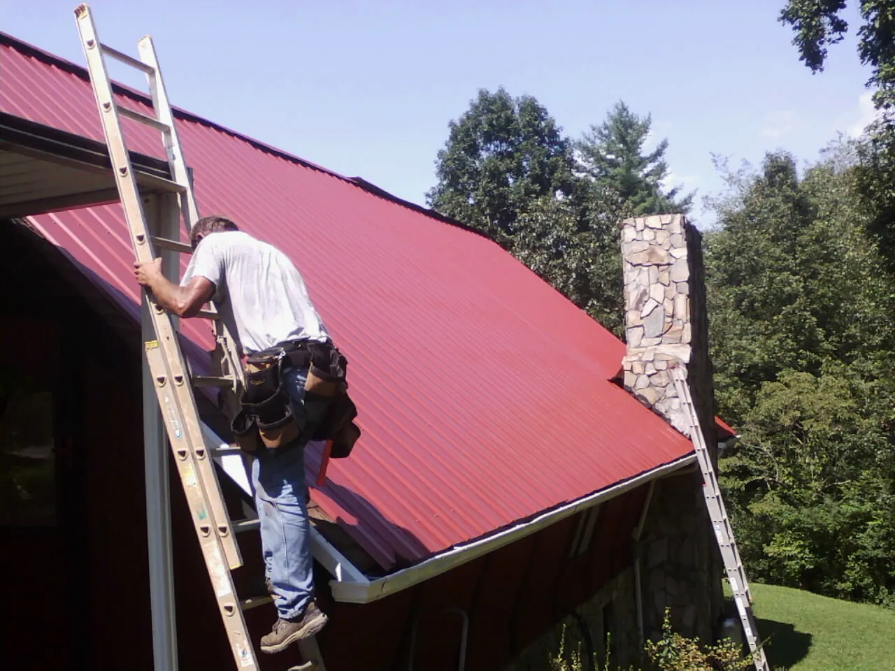 Roofer on a ladder beside a red metal roof and stone chimney — Soffit & Fascia Repair by Roofing Brothers in Michigan Center